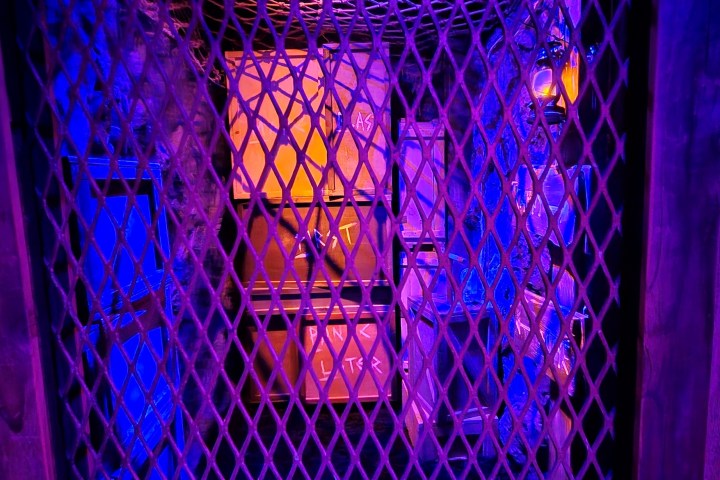 Metal grate with purple lighting revealing stacked crates and a mannequin's face.