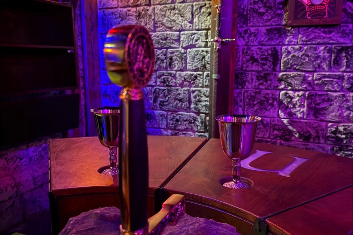 Sword in stone with goblets on wooden tables, brick wall background, purple lighting.
